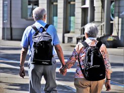 Back view of an elderly couple holding hands and carrying rucksacks while walking down a street